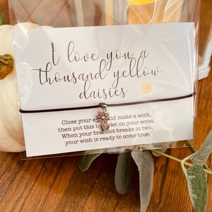 May include: A white card with the text "I love you a thousand yellow daisies" and a small daisy charm attached to a brown string bracelet. The card also includes instructions for making a wish with the bracelet.