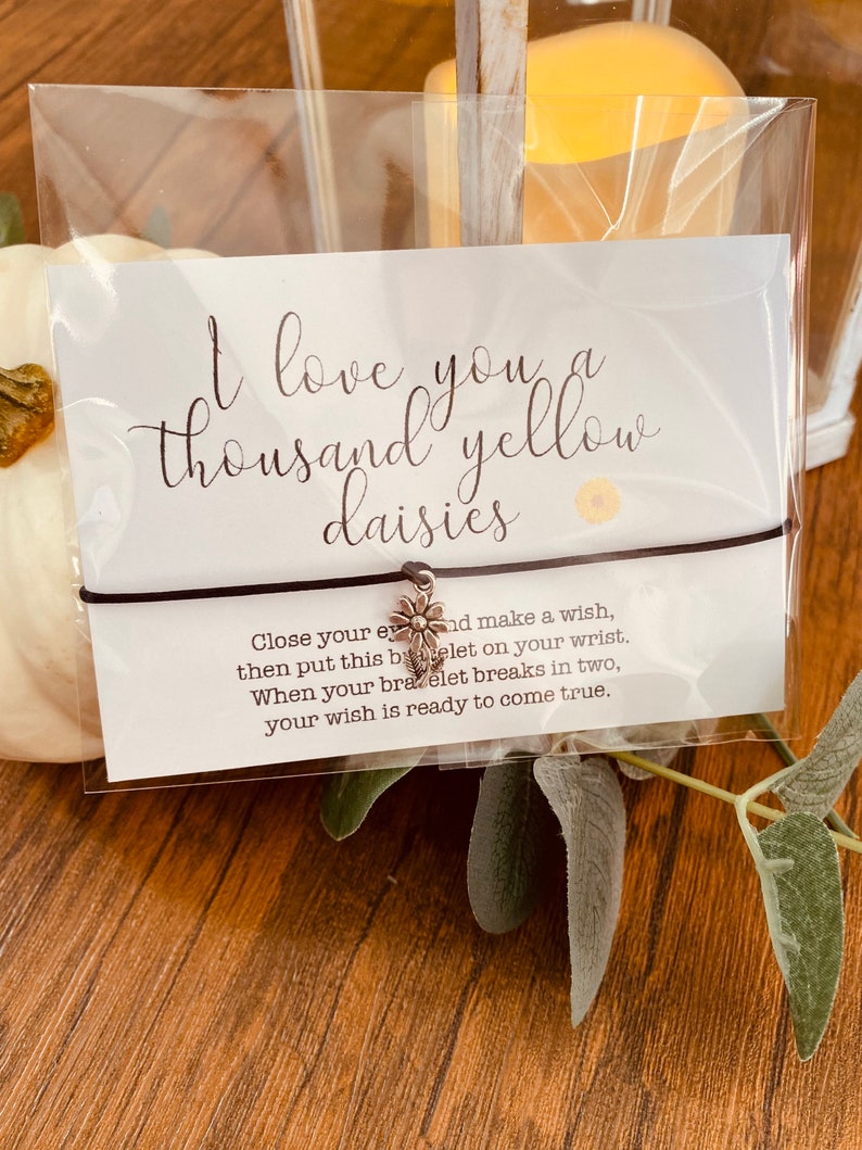 May include: A white card with the text "I love you a thousand yellow daisies" and a small daisy charm attached to a brown string bracelet. The card also includes instructions for making a wish with the bracelet.