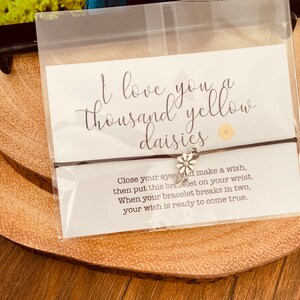 May include: A brown string bracelet with a silver daisy charm. The bracelet is packaged with a card that reads "I love you a thousand yellow daisies." The card also includes instructions for making a wish: "Close your eyes and make a wish, then put this bracelet on your wrist. When your bracelet breaks in two, your wish is ready to come true."