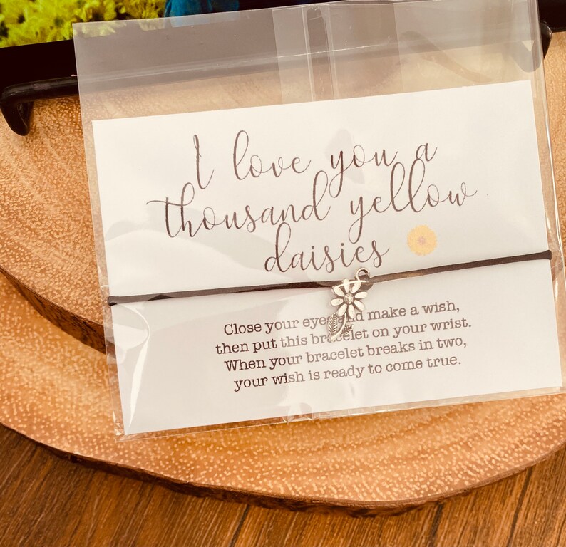 May include: A brown string bracelet with a silver daisy charm. The bracelet is packaged with a card that reads "I love you a thousand yellow daisies." The card also includes instructions for making a wish: "Close your eyes and make a wish, then put this bracelet on your wrist. When your bracelet breaks in two, your wish is ready to come true."