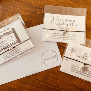 May include: Three wish bracelets with different charms and sayings. The first bracelet has a silver star charm and the saying "I'd rather be in Stars Hollow Founded 1779". The second bracelet has a silver daisy charm and the saying "I love you a thousand yellow daisies". The third bracelet has a silver snowflake charm and the saying "I smell snow".