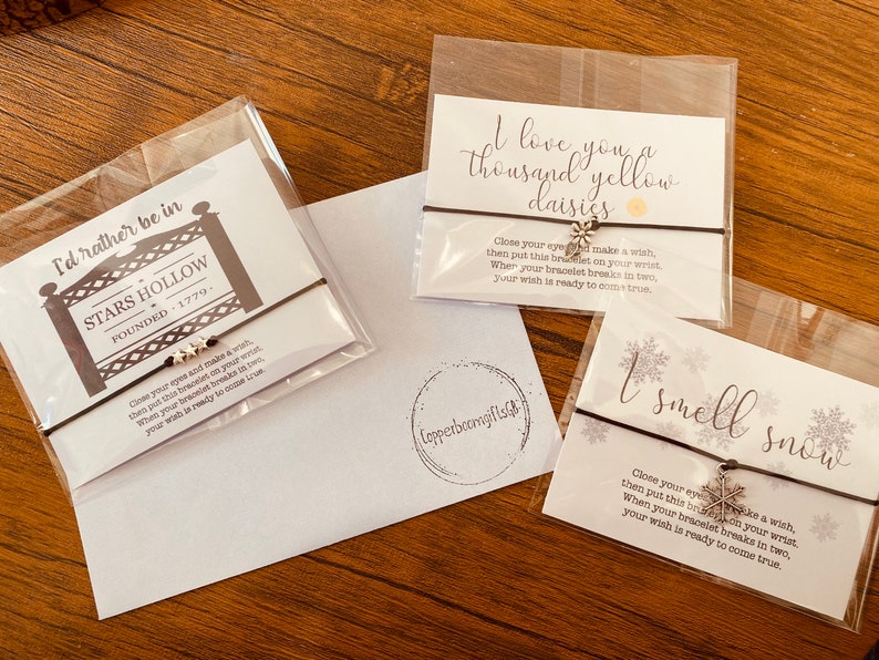 May include: Three wish bracelets with different charms and sayings. The first bracelet has a silver star charm and the saying "I'd rather be in Stars Hollow Founded 1779". The second bracelet has a silver daisy charm and the saying "I love you a thousand yellow daisies". The third bracelet has a silver snowflake charm and the saying "I smell snow".