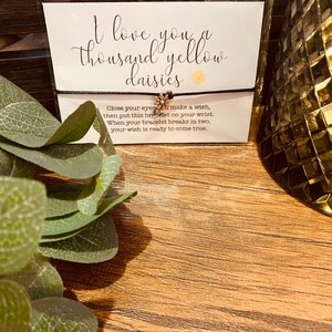May include: A black string bracelet with a gold daisy charm. The bracelet is packaged with a card that reads "I love you a thousand yellow daisies" and instructions to make a wish when the bracelet breaks.