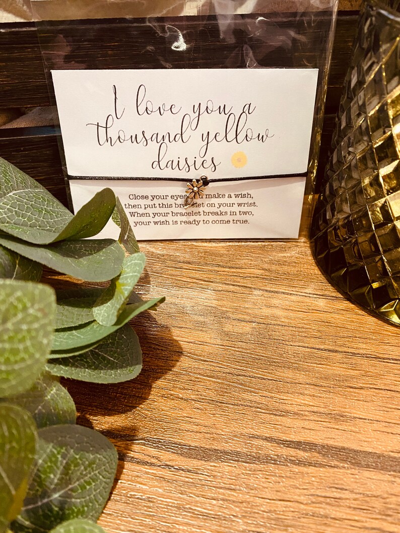 May include: A black string bracelet with a gold daisy charm. The bracelet is packaged with a card that reads "I love you a thousand yellow daisies" and instructions to make a wish when the bracelet breaks.