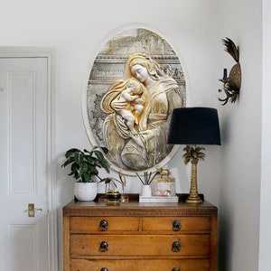 May include: A white oval wall decor featuring a raised relief sculpture of a mother and child. The mother is holding the child in her arms and they are both looking down. The sculpture is set against a backdrop of a city scene.