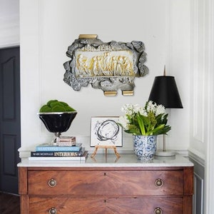 May include: A white and gold wall sculpture depicting a procession of figures in relief. The sculpture is mounted on a white wall above a wooden dresser with two drawers. The dresser is topped with a marble surface and decorated with books, a vase of white flowers, and a small easel with a black and white abstract painting.