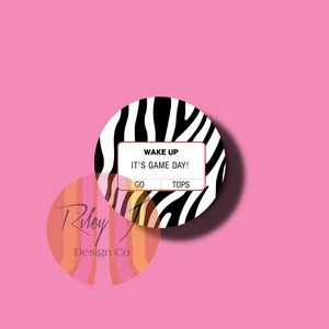 May include: Two round stickers on a pink background. The top sticker has a zebra print border with the words "WAKE UP IT'S GAME DAY!" and the words "GO TOPS" in a white box. The bottom sticker has the words "Riley Design Co" in a colorful design.