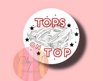 May include: A white circular sticker featuring a black and white illustration of a football stadium. The words "TOPS ON TOP" are written in red lettering around the stadium.