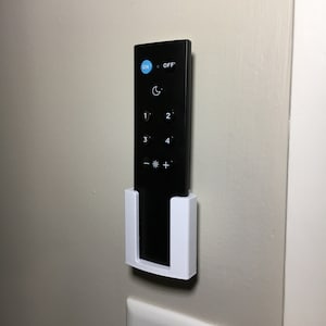May include: A white wall-mounted remote control holder with a black remote control inside. The remote has buttons labeled "ON", "OFF", "1", "2", "3", "4", "*" and "+".
