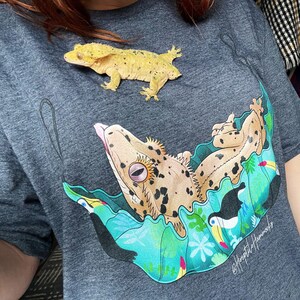 May include: A heather gray t-shirt featuring a gecko graphic with a toucan and tropical leaves. A live gecko rests on the shirt. The graphic includes the text "#HeptileHammocks".