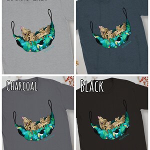 May include: Four t-shirts in sports grey, dark heather, charcoal, and black. Each shirt features a graphic of a gecko relaxing in a hammock made of tropical leaves and toucans. The text "SPORTS GREY", "DARK HEATHER", "CHARCOAL", and "BLACK" are displayed above each shirt.