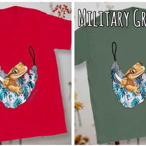 May include: A red and a military green t-shirt with a cartoon illustration of a crested gecko in a hammock. The gecko is light brown with a white belly and is sitting in a blue and white hammock with green leaves and red berries.