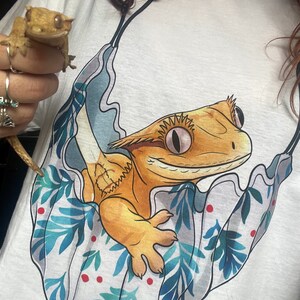May include: White t-shirt with a cartoon illustration of a gecko in a hammock with tropical leaves and red dots. The gecko is orange and yellow with a white belly and pink eyes. The text "@happy.reptile.momma" is at the bottom of the design.