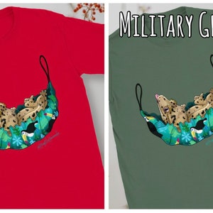 May include: Red t-shirt featuring a whimsical illustration of a leopard-like creature lounging in a hammock adorned with tropical flowers and toucans. The design is set against a solid red background. The text "RED" is at the top.