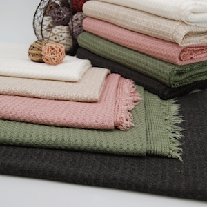 May include: A stack of waffle weave fabric in various colors, including white, beige, pink, green, and gray. The fabric has a textured waffle weave pattern and is folded neatly.