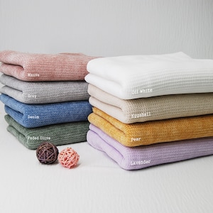May include: A stack of waffle weave fabric swatches in various colors, including mauve, gray, denim, faded olive, eggshell, pear, and lavender. The fabric is folded and stacked neatly, with the color names written on each piece.
