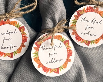 May include: Three gray napkins tied with twine and decorated with round paper tags. Each tag features a fall leaf design and the text "thankful for" followed by a name: Hunter, Willow, and Asher.