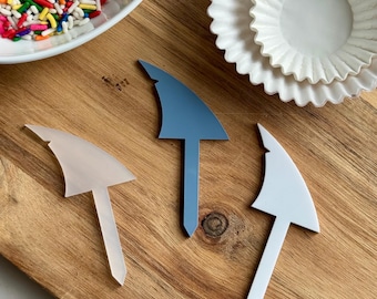 May include: A wooden cutting board holds a white oval bowl filled with colorful sprinkles, white cupcake liners, and three shark fin-shaped cake toppers in light pink, blue, and white. The toppers have a pointed base.