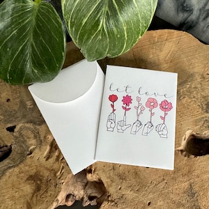 May include: A white card with the text "let love" in black calligraphy. Five pink and red flowers are drawn with black outlines, each held by a hand in American Sign Language. The card is in a white envelope.