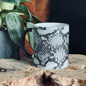 May include: A black and white snake skin patterned ceramic mug with a black handle. The mug is sitting on a piece of wood.