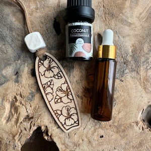 May include: A wooden surfboard-shaped ornament with a floral design, a white wooden bead, and two bottles of essential oil. One bottle is labeled "Coconut Premium Essential Oil" and contains 5 ml (0.17 fl oz).