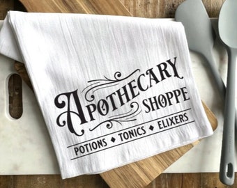 May include: White kitchen towel with black text that reads "Apothecary Shoppe" with the words "Potions, Tonics, Elixers" underneath.