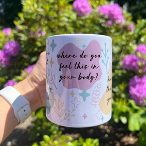 Op de afbeelding: Witte keramische mok met de tekst "where do you feel this in your body?" in zwart schrift. De mok heeft pastelkleurige vormen en botanische illustraties. De mok wordt vastgehouden voor een achtergrond van groen gebladerte en paarse bloemen.
