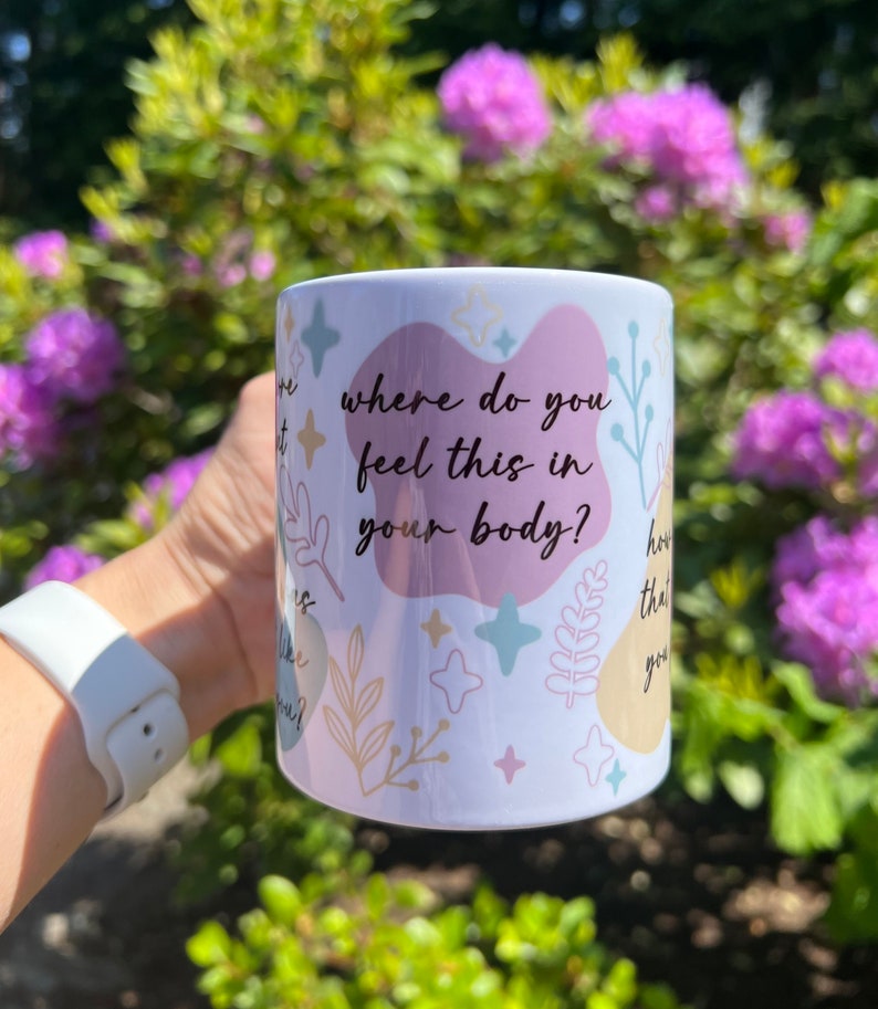 Op de afbeelding: Witte keramische mok met de tekst "where do you feel this in your body?" in zwart schrift. De mok heeft pastelkleurige vormen en botanische illustraties. De mok wordt vastgehouden voor een achtergrond van groen gebladerte en paarse bloemen.