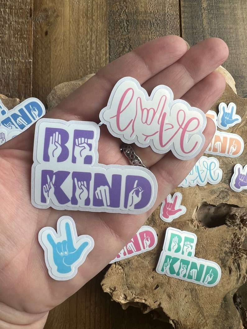 Puede incluir: Una mano sosteniendo varias pegatinas con las palabras "Be Kind" y "Love" escritas en lenguaje de se&ntilde;as. Las pegatinas son de varios colores, incluyendo morado, azul, verde y rosa.