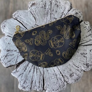 May include: A black fabric pouch with a gold zipper closure. The pouch is decorated with a repeating pattern of gold skulls, snakes, and geometric shapes.