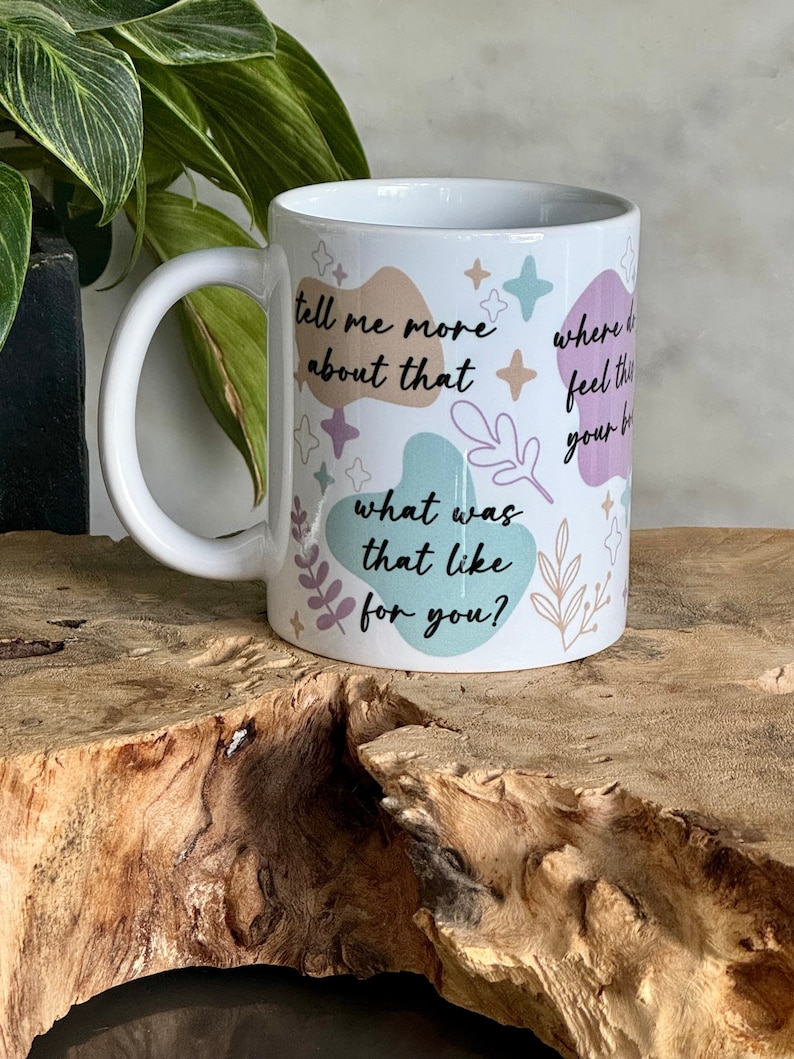 Op de afbeelding: Witte keramische mok met een gebogen handvat, versierd met pastelkleurige vormen en botanische illustraties. De mok bevat de zinnen "tell me more about that", "what was that like for you?" en "where do you feel this in your body?"