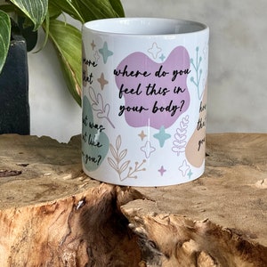 Op de afbeelding: Witte keramische mok met de tekst "where do you feel this in your body?" in het zwart. De mok heeft pastelkleurige abstracte vormen en botanische illustraties. De mok is cilindrisch en staat op een houten oppervlak.