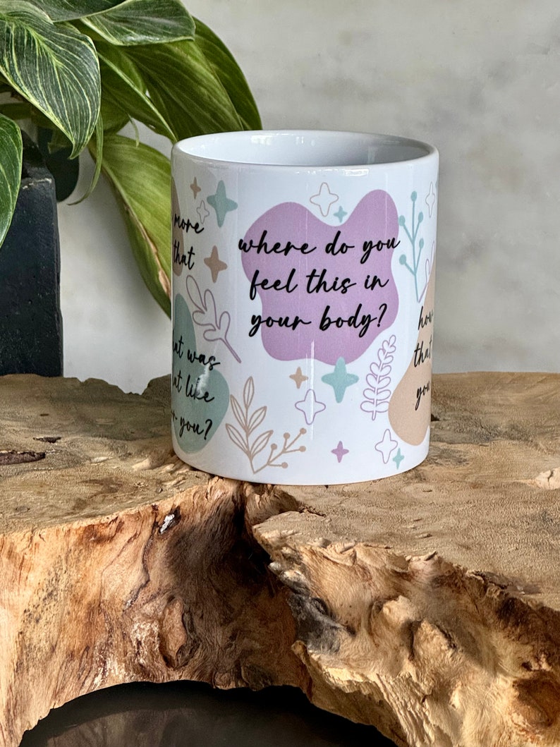 Op de afbeelding: Witte keramische mok met de tekst "where do you feel this in your body?" in het zwart. De mok heeft pastelkleurige abstracte vormen en botanische illustraties. De mok is cilindrisch en staat op een houten oppervlak.