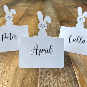 May include: Three white bunny place card holders with the names Peter, April, and Calla written on them.