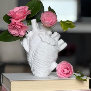 May include: A white ceramic heart-shaped vase with pink camellia flowers arranged inside. The vase is sitting on top of a stack of two books, one beige and one pink.