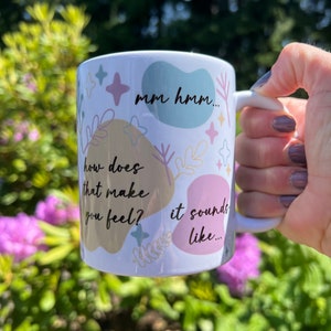 Op de afbeelding: Witte keramische mok met pastelkleurige vormen, sterren en bloemmotieven. De mok heeft de zinnen "mm hmm...", "how does that make you feel?" en "it sounds like..." in zwarte tekst. De mok wordt vastgehouden tegen een wazige groene en paarse achtergrond.