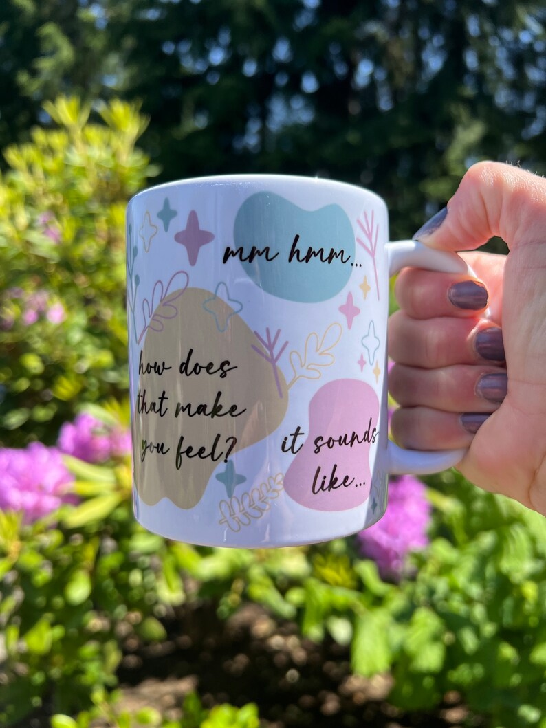 Op de afbeelding: Witte keramische mok met pastelkleurige vormen, sterren en bloemmotieven. De mok heeft de zinnen "mm hmm...", "how does that make you feel?" en "it sounds like..." in zwarte tekst. De mok wordt vastgehouden tegen een wazige groene en paarse achtergrond.