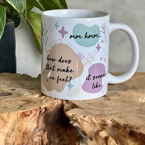 Op de afbeelding: Witte keramische mok met de tekst "mm hmm... how does that make you feel? it sounds like..." in het zwart. De mok heeft een oor en is versierd met pastelkleurige vormen en bloemmotieven.