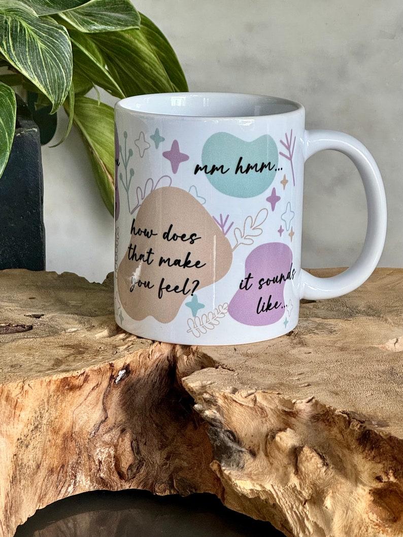 Op de afbeelding: Witte keramische mok met de tekst "mm hmm... how does that make you feel? it sounds like..." in het zwart. De mok heeft een oor en is versierd met pastelkleurige vormen en bloemmotieven.