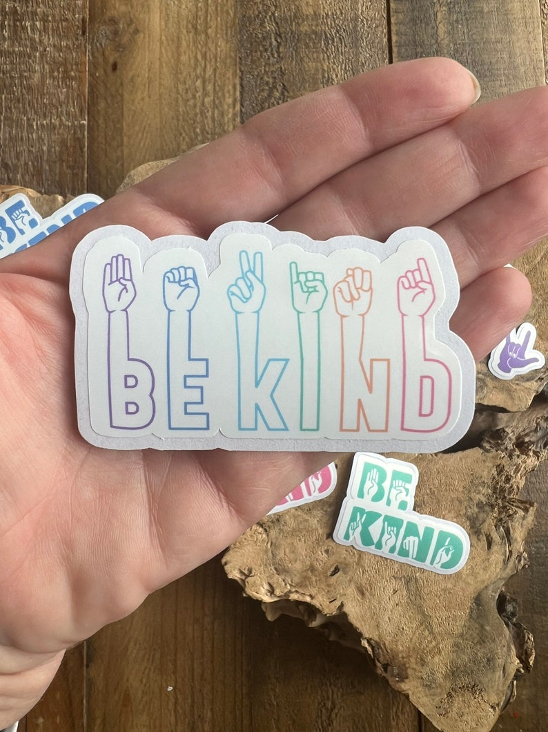 Puede incluir: Una pegatina blanca con un dise&ntilde;o degradado arco&iacute;ris. La pegatina presenta la palabra "BE KIND" en lenguaje de se&ntilde;as. La pegatina est&aacute; sostenida en una mano.