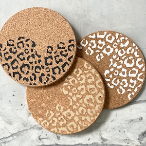 May include: Three round cork coasters with a leopard print design. The coasters are in black, white, and beige.