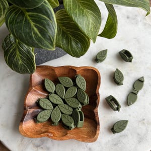 May include: A wooden bowl filled with green leaf-shaped hair clips. Several additional clips are scattered on a white surface.