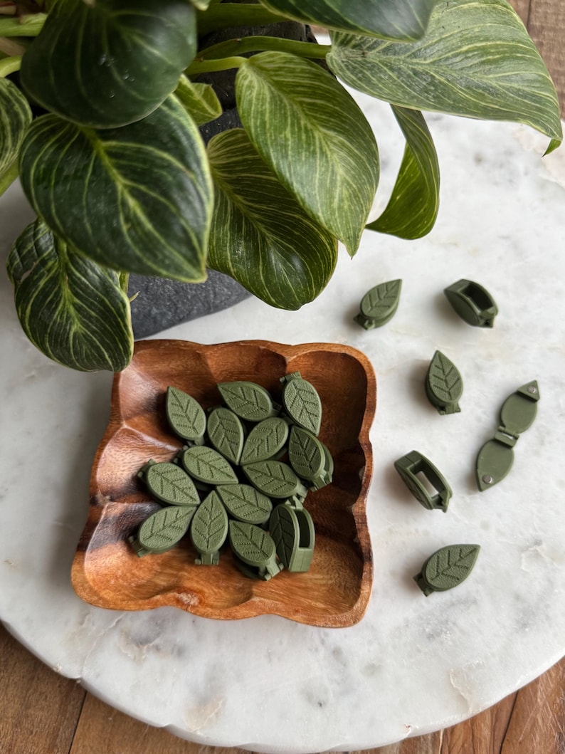 May include: A wooden bowl filled with green leaf-shaped hair clips. Several additional clips are scattered on a white surface.