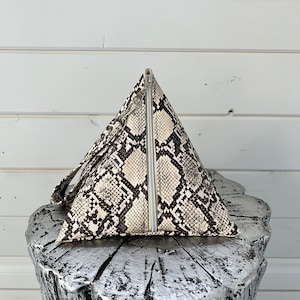 May include: A triangular-shaped handbag with a zipper closure and a snakeskin pattern. The bag is made of a faux leather material and has a strap.