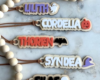 May include: Wooden Halloween-themed name tags with names like "Lilith," "Cordelia," "Thoren," "Syndea," and "Silas." Each tag features a unique Halloween icon, such as a ghost, pumpkin, bat, witch hat, and skull. The tags are strung on a wooden bead garland.
