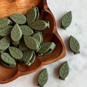 May include: A bowl filled with green leaf-shaped clips, with additional clips scattered on a white surface. The clips have a textured surface and a detailed leaf design.