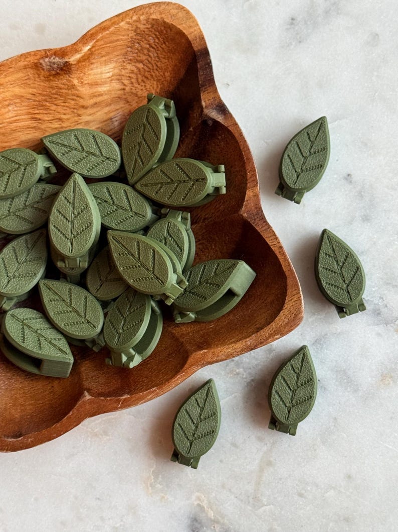 May include: A bowl filled with green leaf-shaped clips, with additional clips scattered on a white surface. The clips have a textured surface and a detailed leaf design.