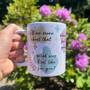 Op de afbeelding: Witte keramische mok met een handvat, versierd met pastelkleurige abstracte vormen, sterren en botanische illustraties. De mok heeft de zinnen "tell me more about that", "where feel your" en "what was that like for you?" erop gedrukt.