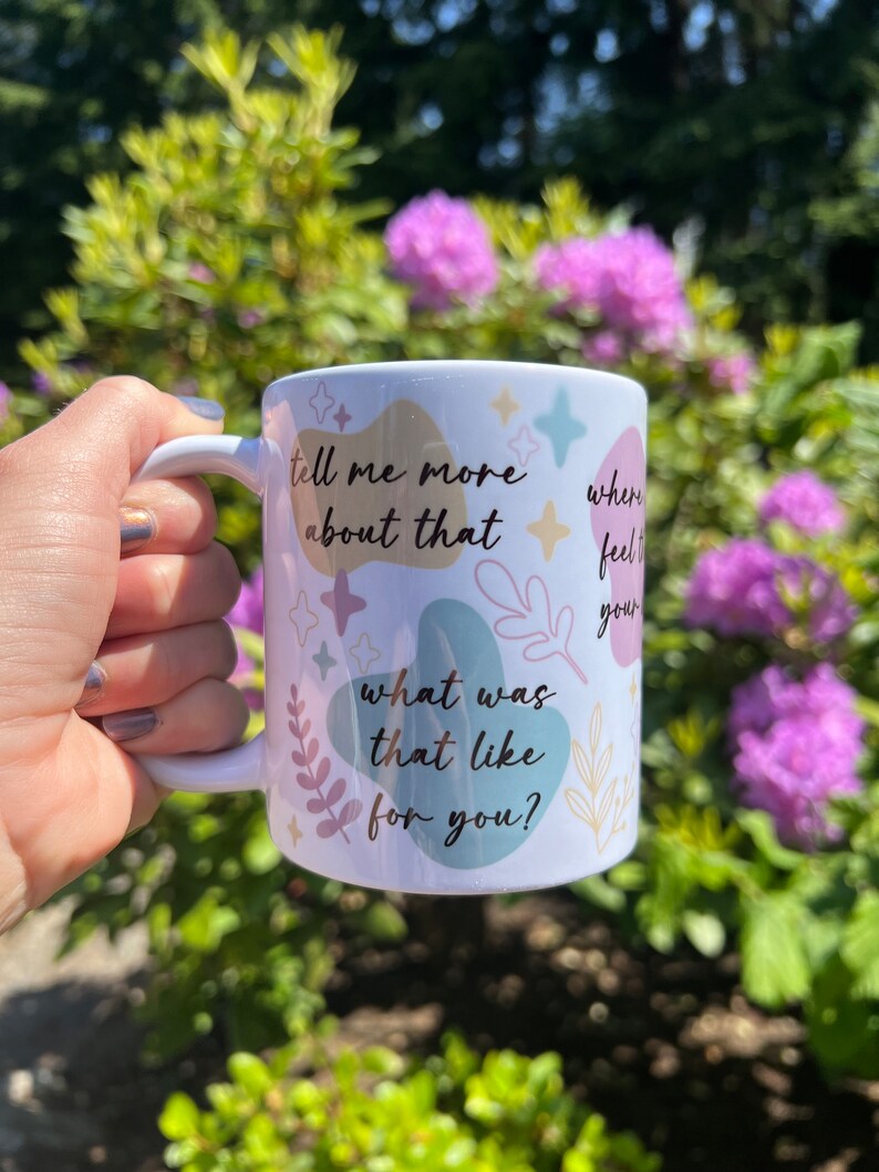 Op de afbeelding: Witte keramische mok met een handvat, versierd met pastelkleurige abstracte vormen, sterren en botanische illustraties. De mok heeft de zinnen "tell me more about that", "where feel your" en "what was that like for you?" erop gedrukt.