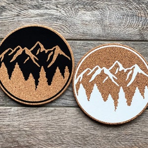 May include: Two round cork coasters with a mountain range and pine trees design. One coaster has a black mountain range and trees, the other has a white mountain range and trees.